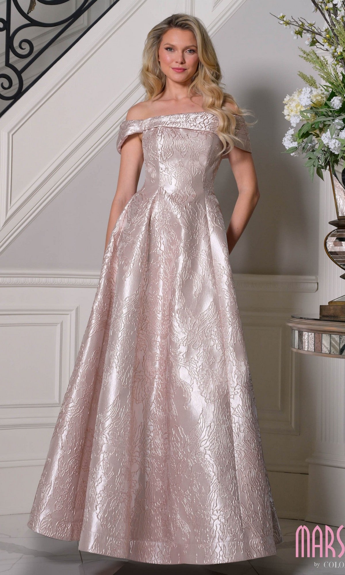 A woman stands indoors wearing the Marsoni MV1395 long formal dress in blush, featuring an off-the-shoulder floor-length jacquard with a floral pattern—an elegant choice for a mother-of-the-bride look.